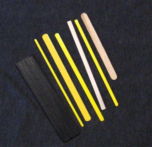 An assortment of mesh sticks including yellow plastic ones, bare wood ones, one of which is a popsicle stick, and a wide, black-painted wood one.